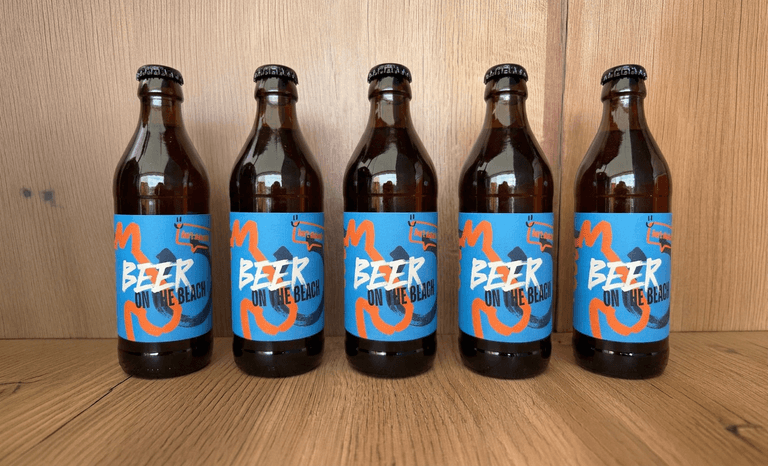 Fünf braune Bierflaschen mit blauen und orangefarbenen Etiketten, auf denen „Beer on the Beach“ steht, stehen in einer Reihe auf einer Holzfläche.