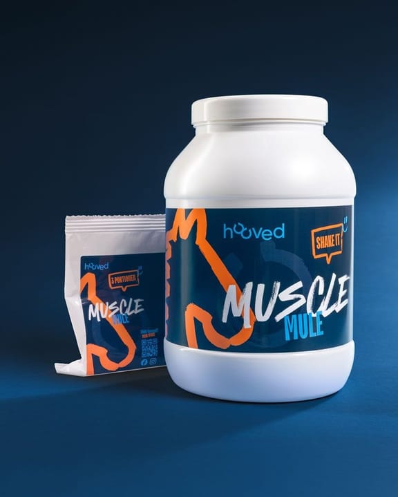 Großer weißer Behälter mit blauer Etikette, die orange und weiße Schrift zeigt "Muscle MULE" und "Shake it". Daneben eine kleinere Packung mit ähnlichem Design.