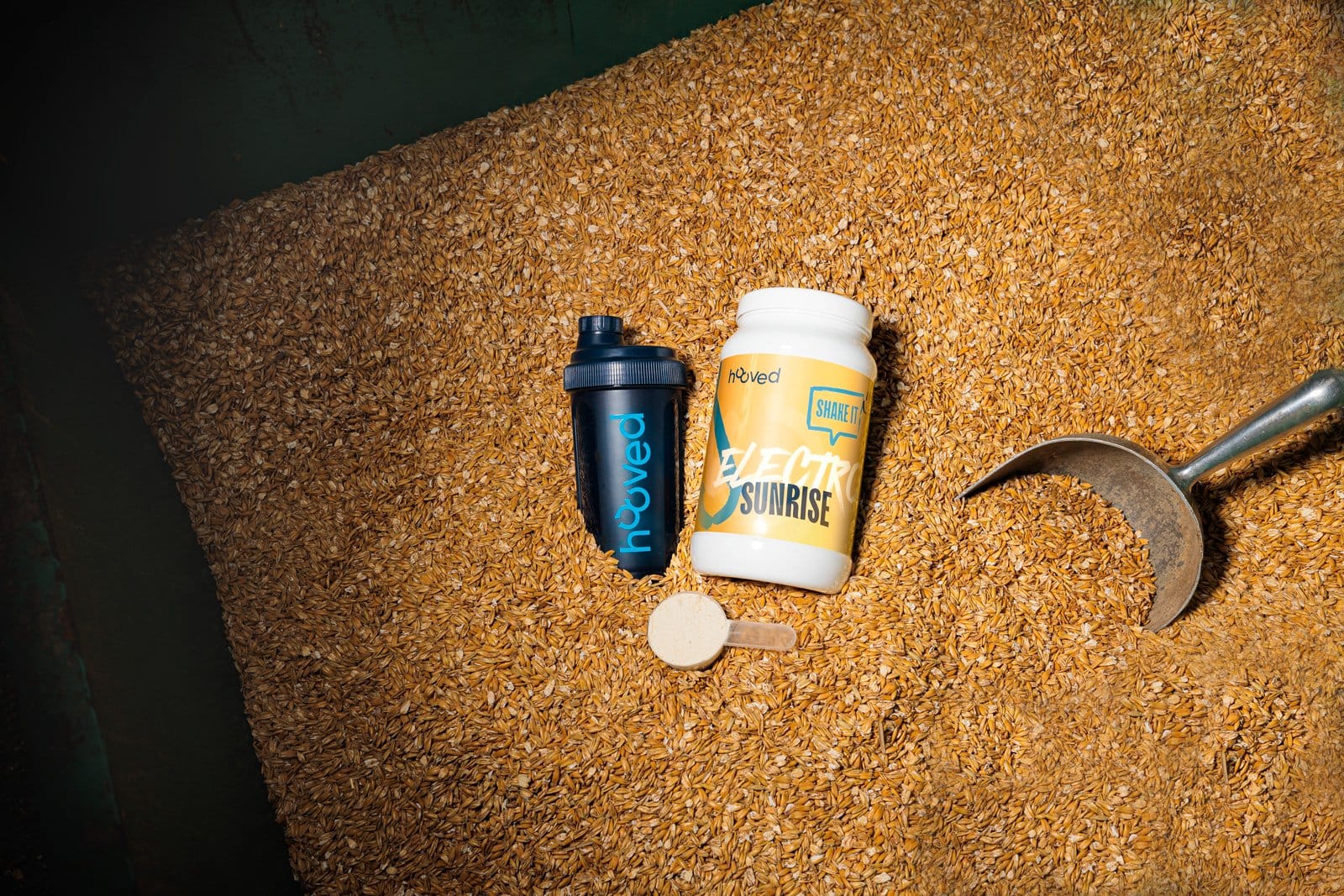 Nahaufnahme von einem schwarzen Messlöffel, einem weißen Proteinpulverbehälter, einem schwarzen Shaker und einem weißen Pulverlöffel auf einer Oberfläche aus braunen Getreidekörnern.
