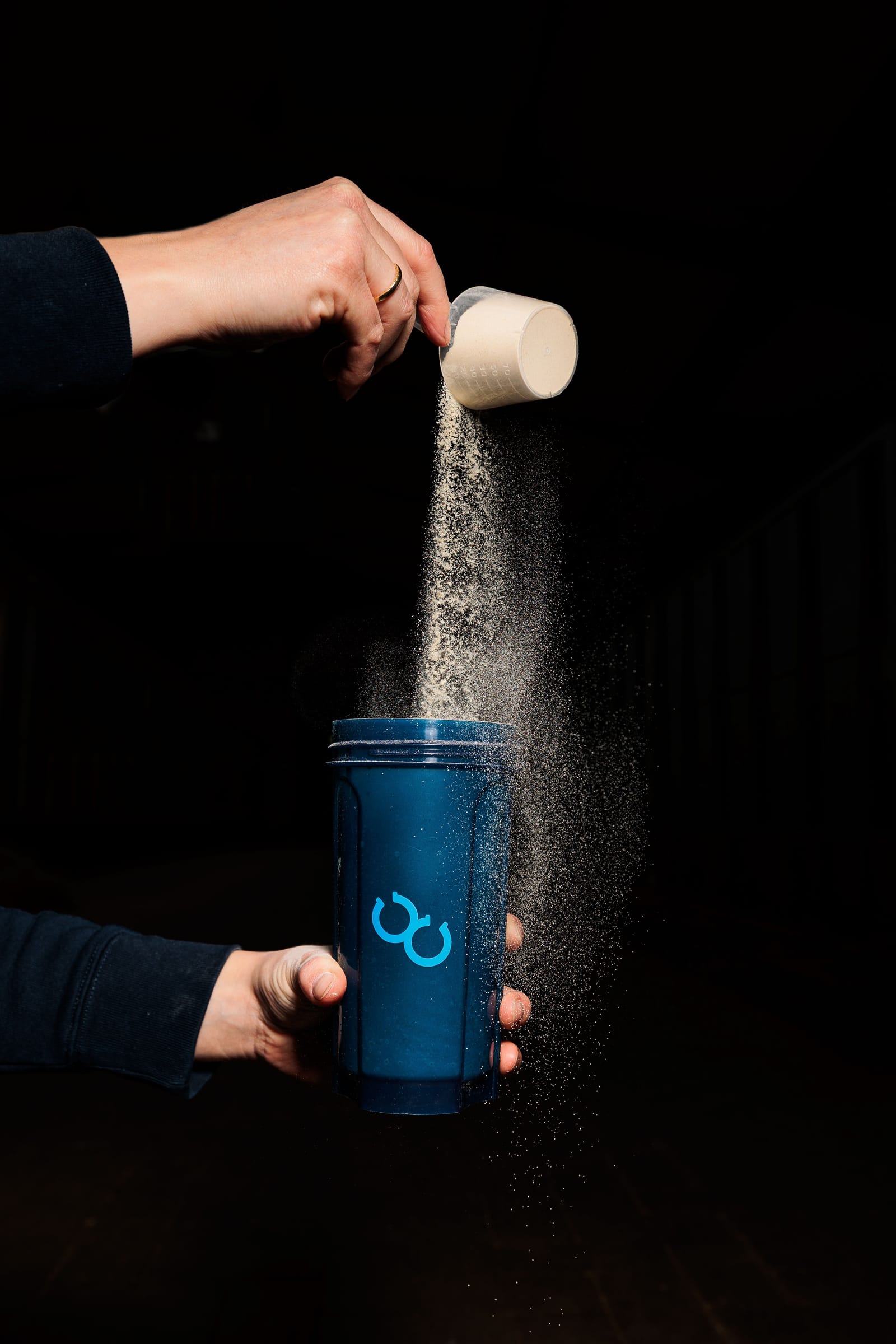 Eine Hand hält eine blaue Thermosflasche, aus der feiner Staub oder Pulver in die Flasche fällt, vor einem schwarzen Hintergrund.